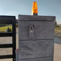 Szary, betonowy słupek bramy z zamontowanym panelem wideodomofonu Eura i pomarańczową lampą ostrzegawczą na szczycie, widok z boku, częściowo widoczna czarna brama.