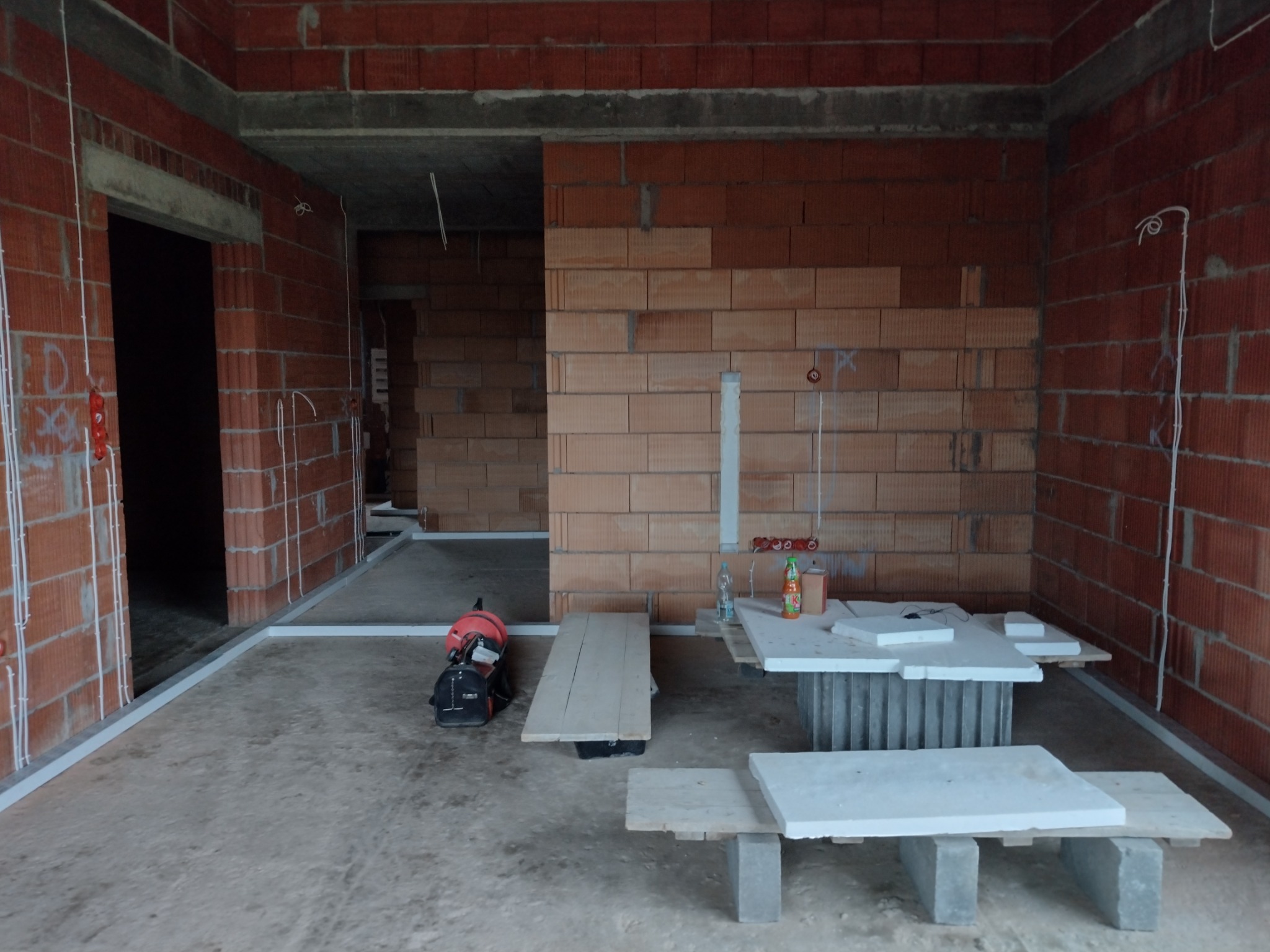 Wnętrze surowego budynku w trakcie prac instalacyjnych: widoczne ściany z czerwonej cegły, białe peszle elektryczne prowadzone wzdłuż ścian, prowizoryczny stół roboczy z materiałami budowlanymi...