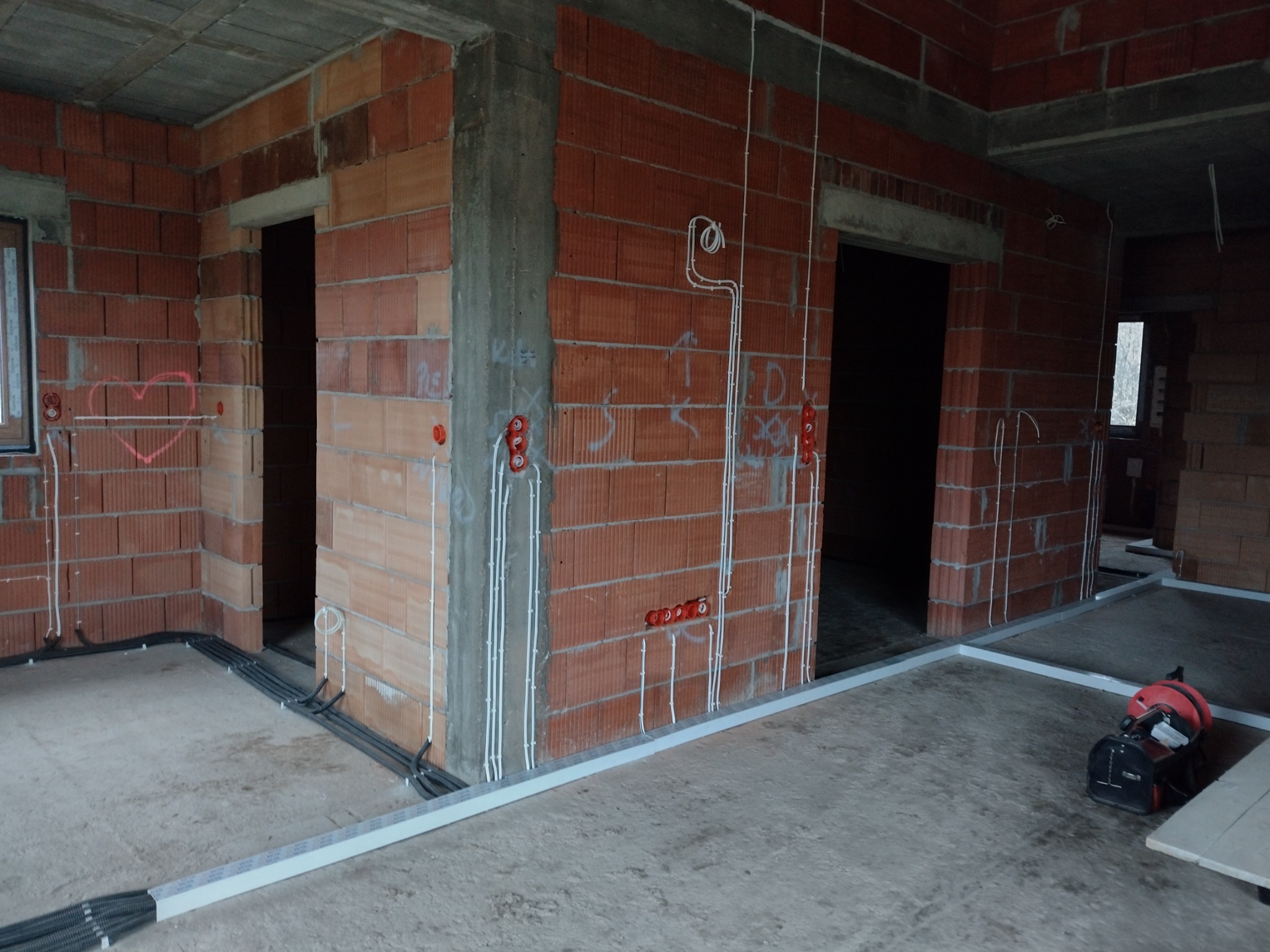 Surowy stan wnętrza budynku w trakcie prac elektrycznych: widoczne przewody elektryczne prowadzone po ścianach z cegły, puszki instalacyjne oraz kable w korytach podłogowych, na podłodze stoi...