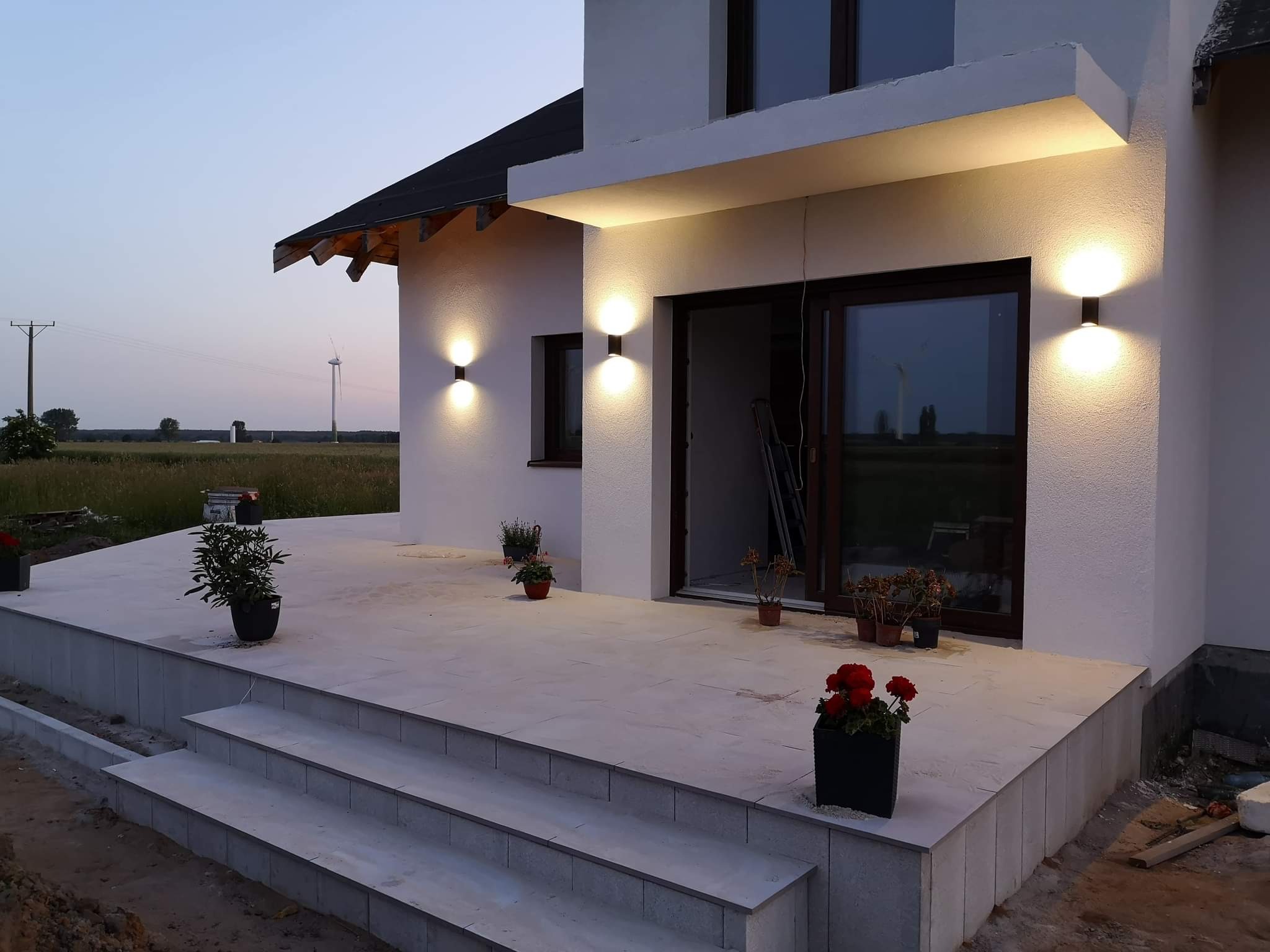 Nowoczesny dom z oświetlonym wejściem i tarasem wyłożonym jasnymi płytami, widoczne lampy elewacyjne, rośliny w donicach i turbina wiatrowa w tle.