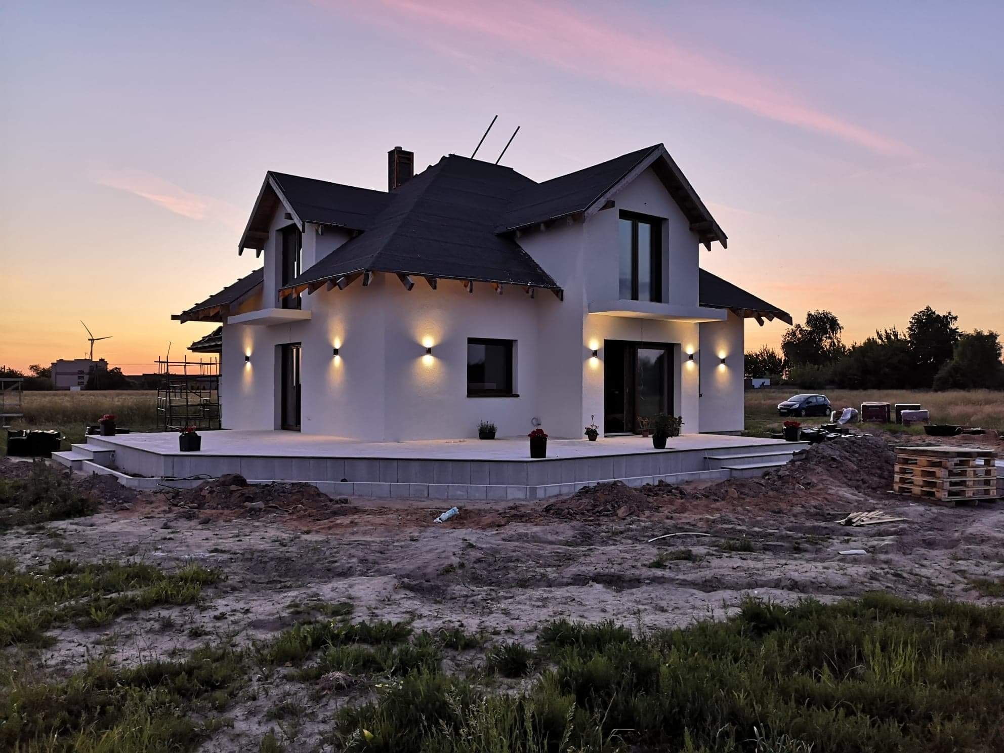 Nowoczesny dom jednorodzinny z białymi ścianami i ciemnym dachem oświetlony zewnętrznymi lampami ściennymi, widoczne elementy instalacji odgromowej na dachu, w tle zachodzące słońce i turbina...