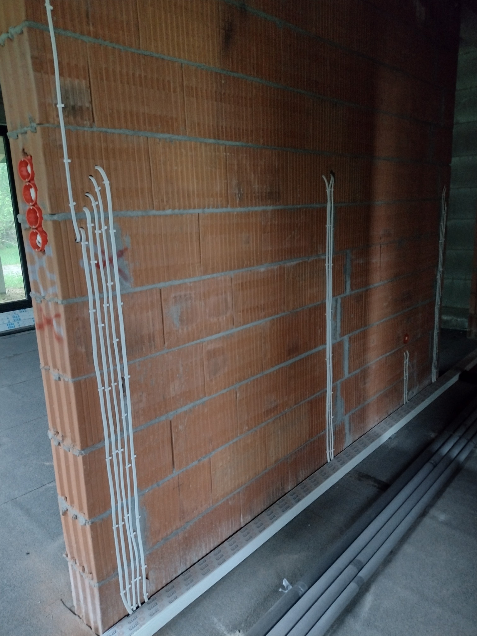 Ściana z czerwonej cegły z pionowo poprowadzonymi białymi przewodami elektrycznymi w peszlach i czerwonymi puszkami instalacyjnymi, widoczne szare rury na podłodze w surowym stanie.