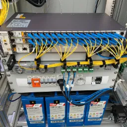 Szafa telekomunikacyjna z panelami krosowniczymi, światłowodami w kolorach niebieskim i żółtym, modułami zasilania i akumulatorami FIAMM 12FIT101, zainstalowana w metalowej ramie.