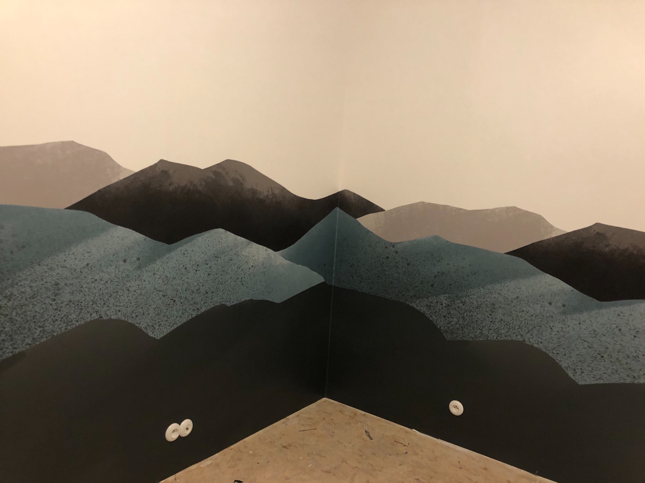 Ściana w rogu pomieszczenia z ręcznie malowanym motywem górskim w odcieniach szarości i błękitu, widoczne gniazdka elektryczne na dole ściany i surowa podłoga.