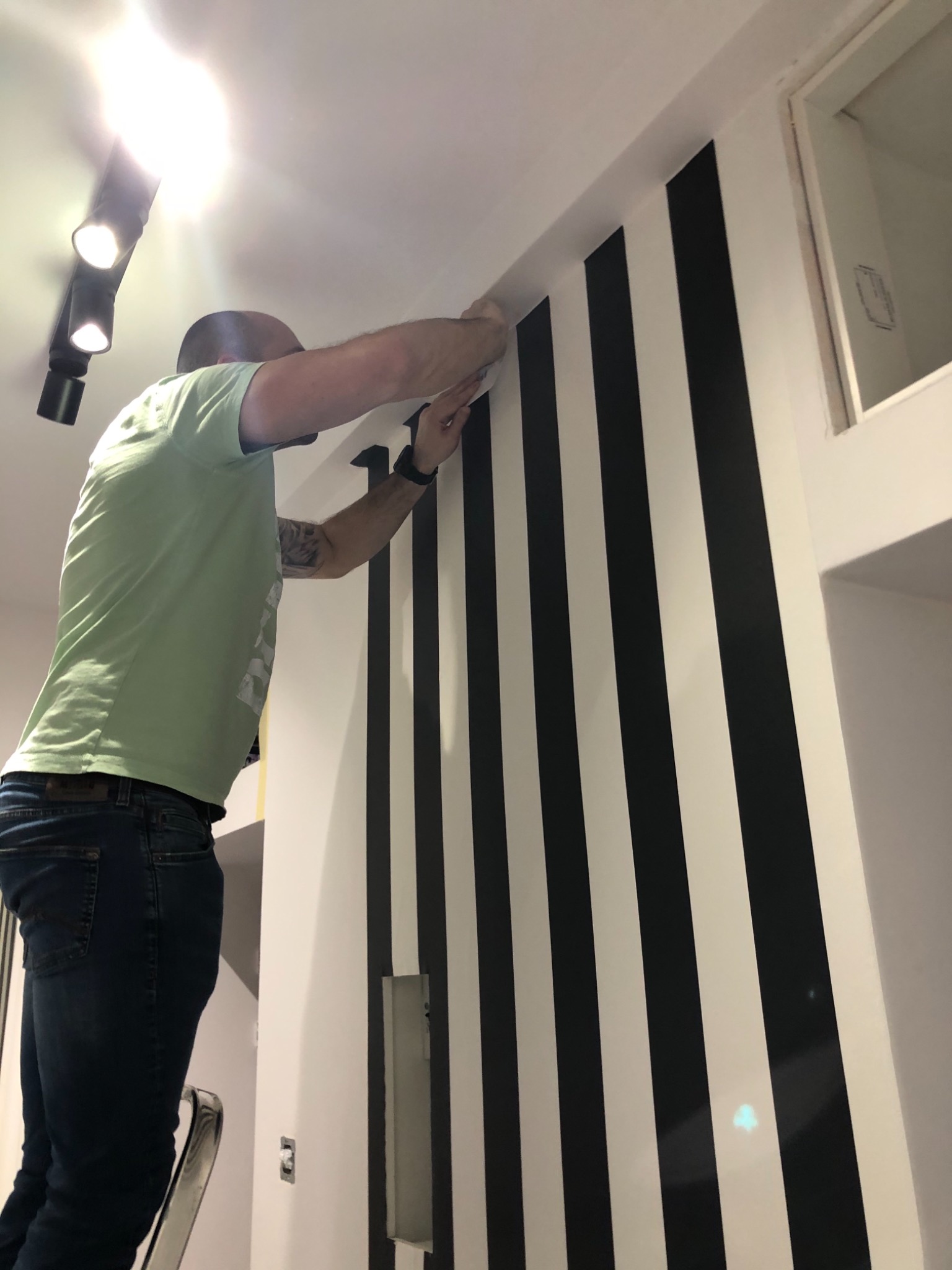Mężczyzna na drabinie, z tatuażem na ramieniu, przykłada czarny pas tapety do ściany w biało-czarne pionowe pasy. Widoczne oświetlenie sufitowe.