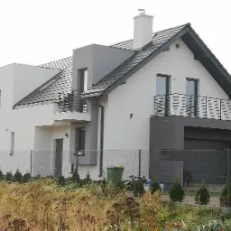 Nowoczesny dom jednorodzinny z elewacją w odcieniach bieli i szarości, widok z zewnątrz na tle brązowej trawy i ogrodzenia z siatki.