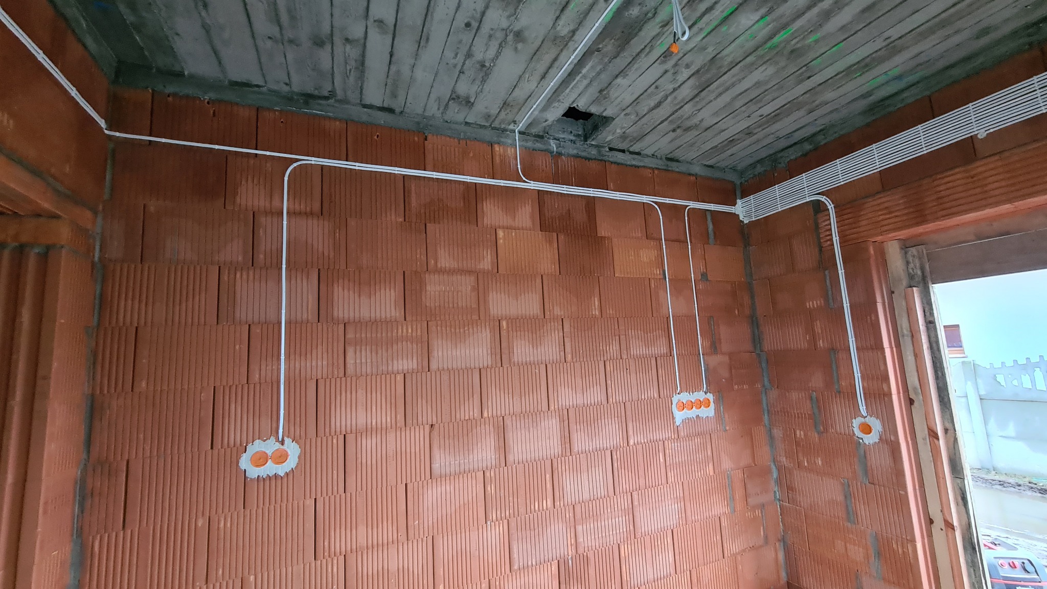 Surowe wnętrze budynku w trakcie budowy z widoczną instalacją elektryczną w białych rurkach PCV, poprowadzoną po czerwonej, ceglanej ścianie i betonowym suficie.