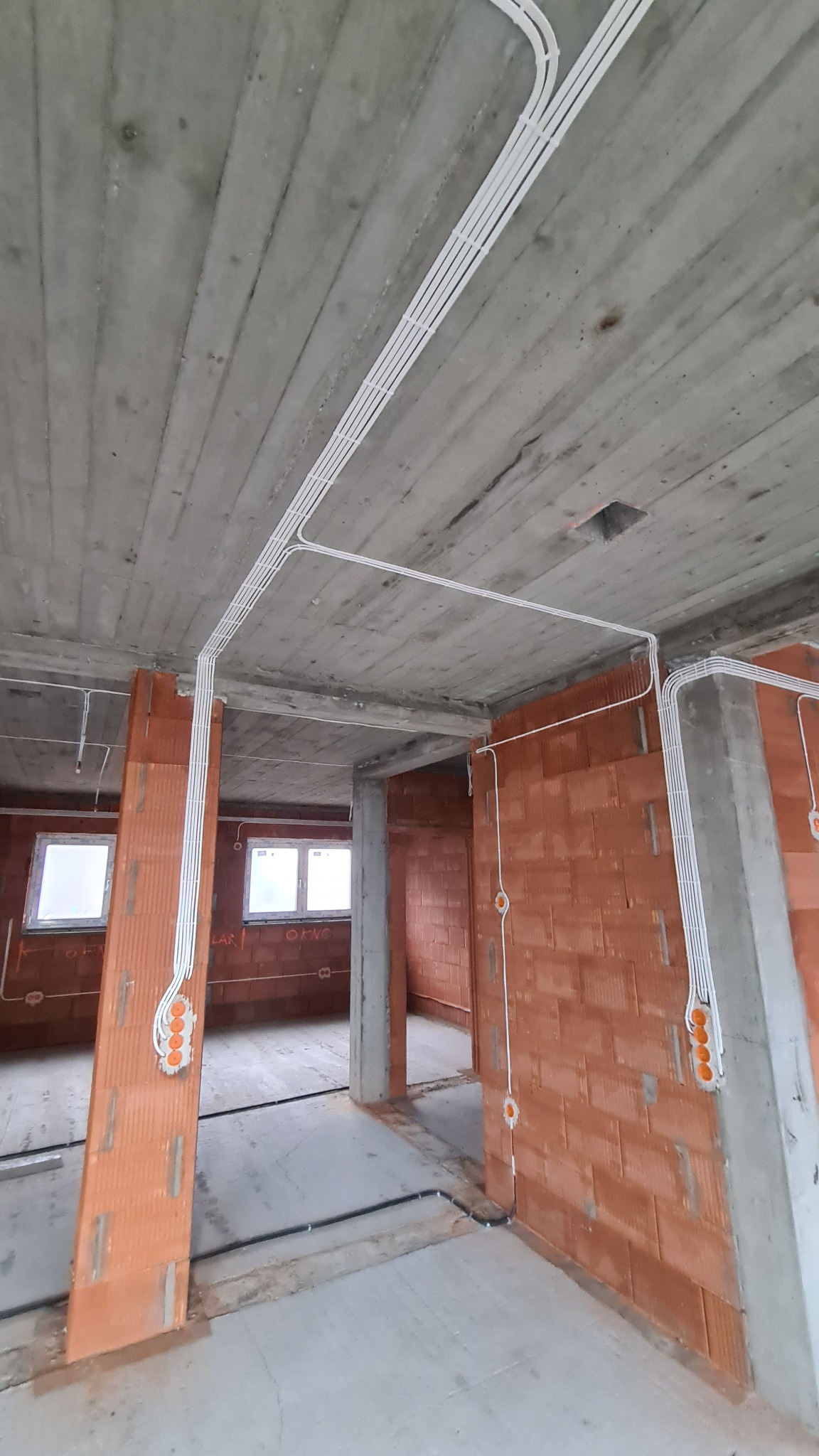 Surowy stan budynku w trakcie prac: widoczne kable elektryczne prowadzone po suficie i ścianach z cegły, puszki instalacyjne na ścianach, betonowy sufit.