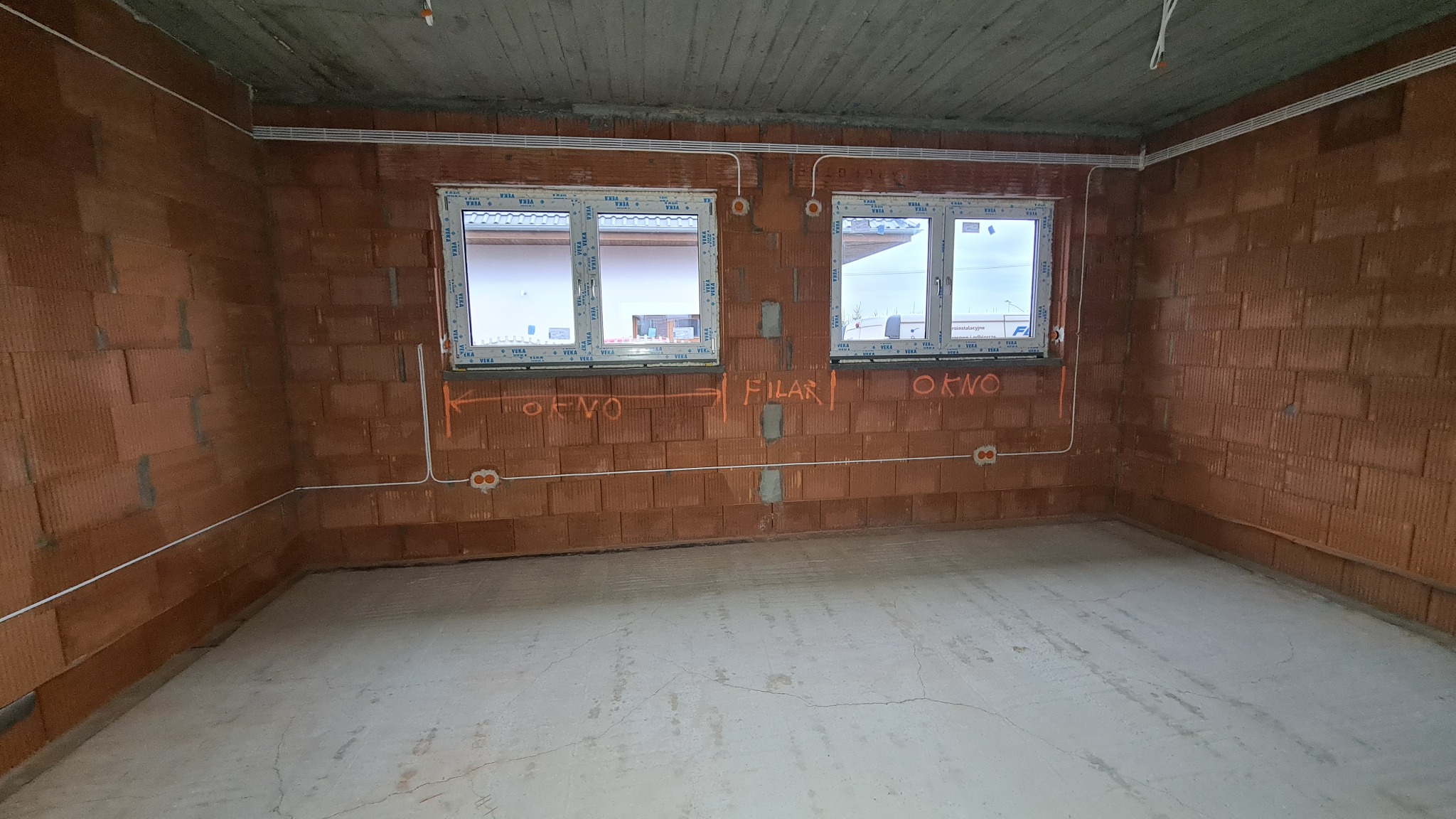 Surowy stan wnętrza z widocznymi przewodami elektrycznymi prowadzonymi po ścianach z czerwonej cegły dziurawki, z dwoma oknami. Na ścianach widoczne pomarańczowe napisy 'OKNO' i 'FILAR'.