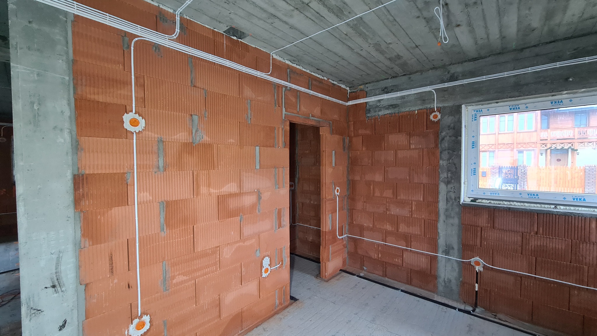 Wnętrze w stanie deweloperskim z widoczną instalacją elektryczną w białych peszlach, prowadzoną po ścianach z cegły ceramicznej i suficie betonowym. Widoczne puszki instalacyjne z pomarańczowymi...