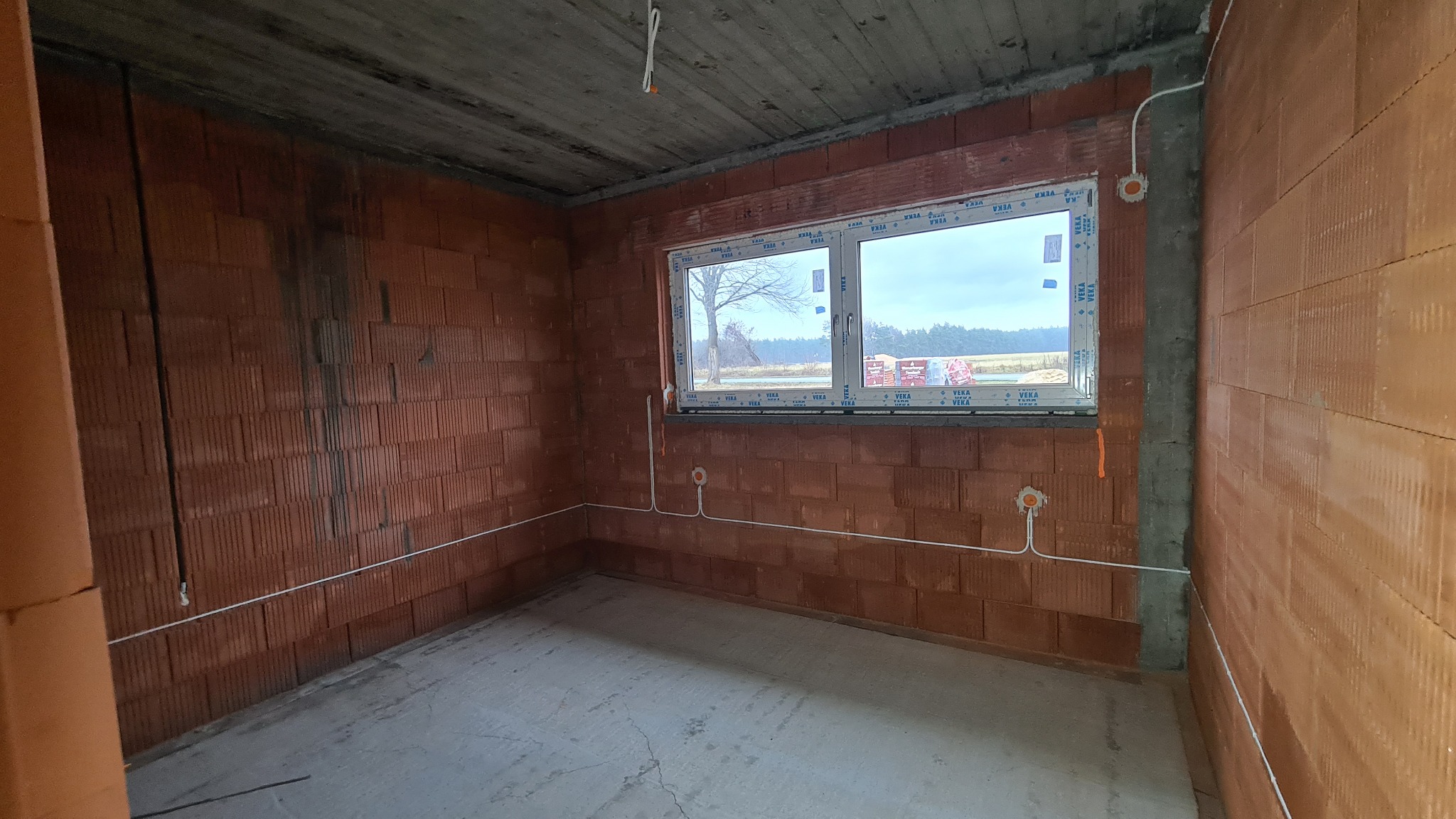 Surowy pokój w trakcie budowy z widocznymi przewodami elektrycznymi wyprowadzonymi ze ściany, okno z widokiem na pole w tle.