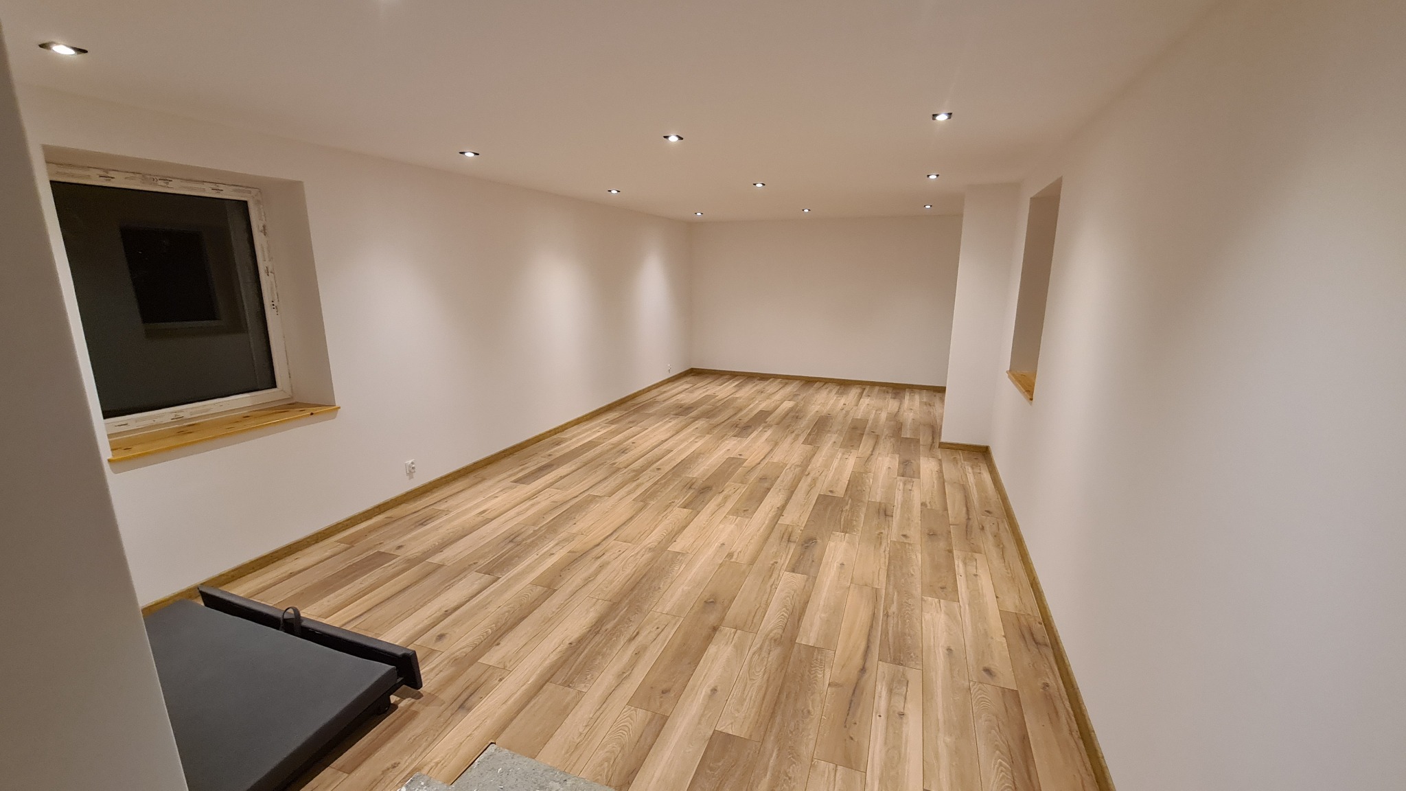 Puste pomieszczenie z panelami podłogowymi w odcieniach drewna, oświetlone punktowymi lampami sufitowymi; okno z drewnianym parapetem po lewej stronie.