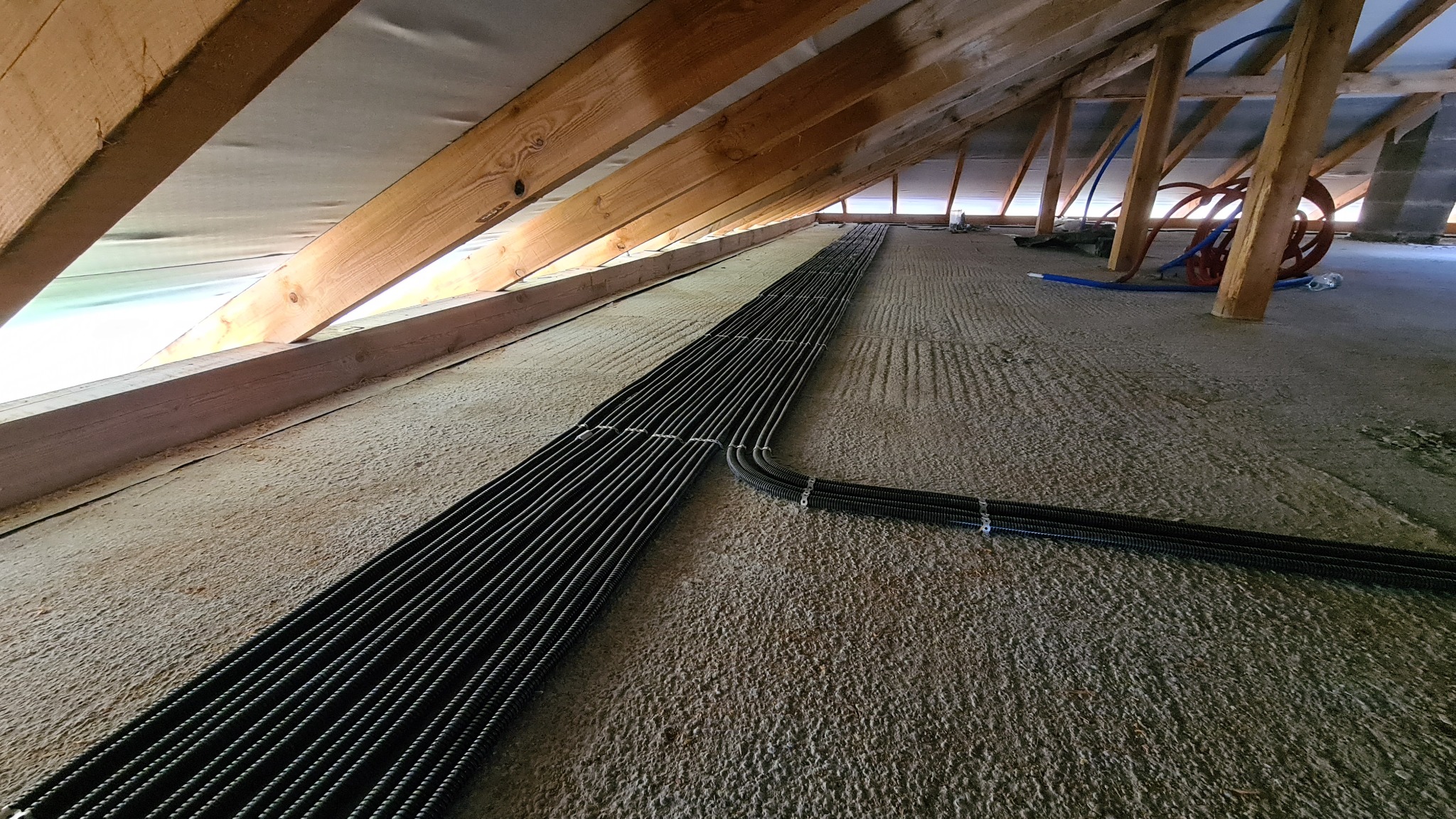 Równolegle ułożone, czarne, karbowane rury instalacyjne na surowym podłożu poddasza, widoczne drewniane elementy konstrukcji dachu.