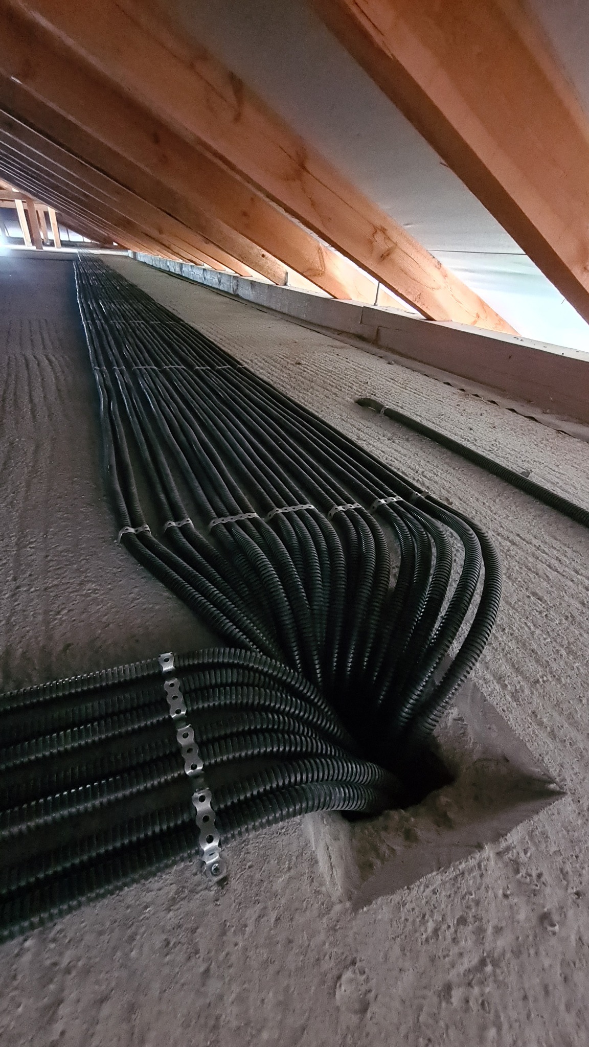 Rzędy czarnych, karbowanych rur prowadzone wzdłuż szarej, betonowej ściany pod drewnianą konstrukcją dachu, zabezpieczone metalowymi uchwytami.
