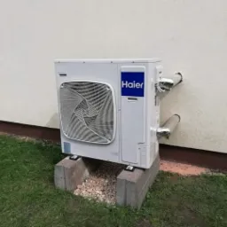 Zewnętrzna jednostka klimatyzacji Haier zamontowana na betonowych blokach przy ścianie budynku, z widocznymi rurami odprowadzającymi skropliny zabezpieczonymi folią.