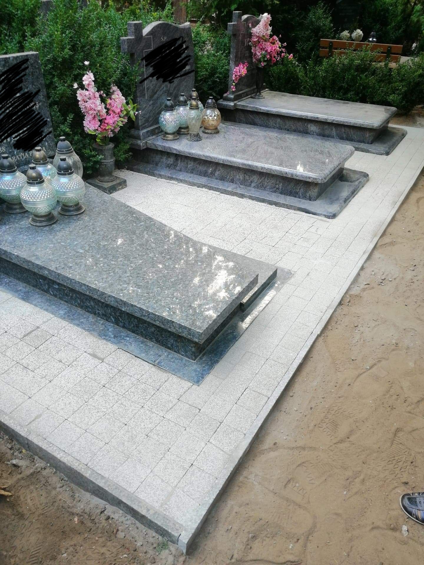 Granitowe nagrobki z zapalonymi lampionami i różowymi kwiatami, otoczone jasną kostką brukową i zielenią cmentarza, widok z góry.