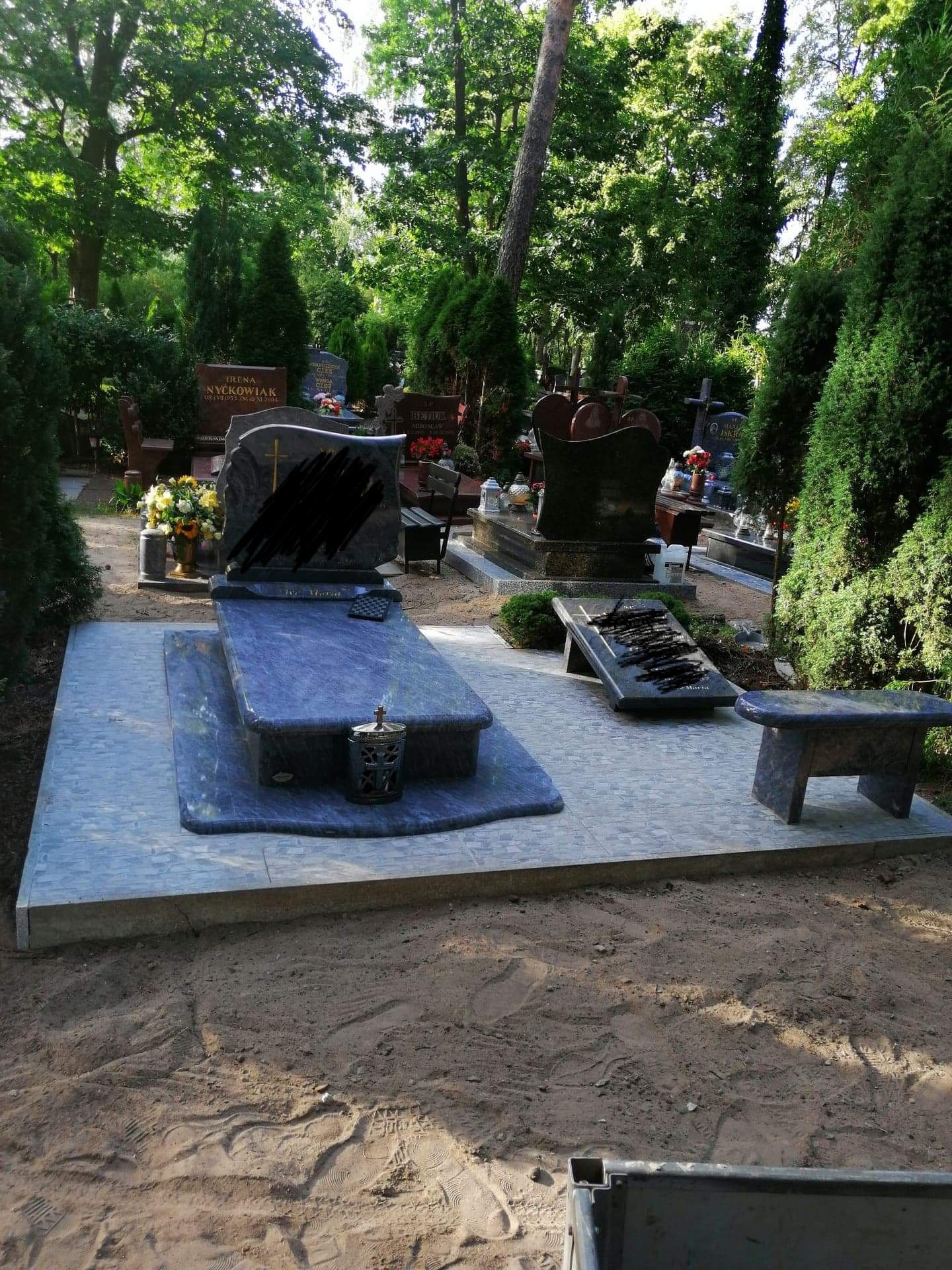 Niebiesko-szary nagrobek z kamienia naturalnego, otoczony kostką brukową, z niewielką kamienną ławką i lampionem, na tle zieleni cmentarza.