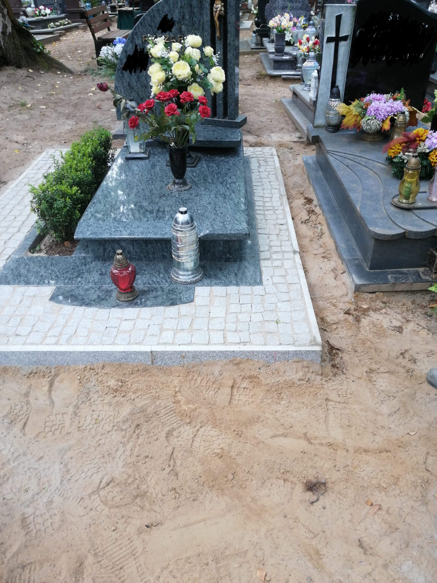 Granitowy pomnik z kwiatami i lampionami, otoczony kostką brukową na cmentarzu, świeżo obsypany piaskiem z widocznymi śladami stóp.