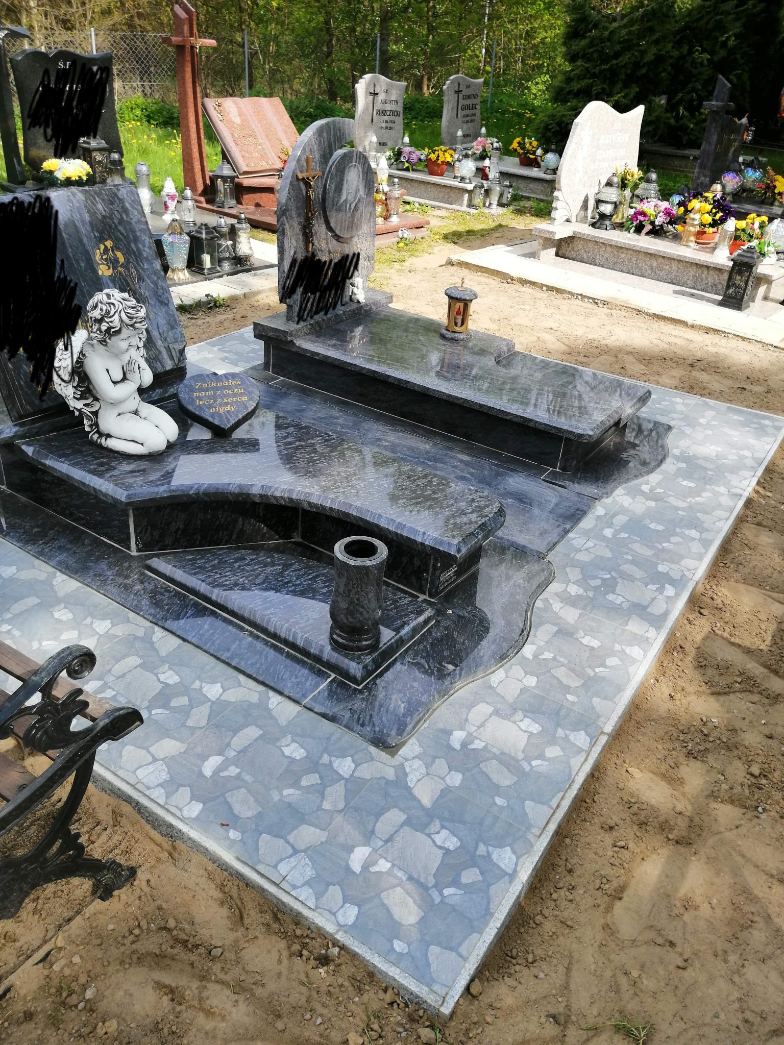 Granitowy nagrobek z marmurową figurką anioła, otoczony płytami chodnikowymi, na cmentarzu w słoneczny dzień.