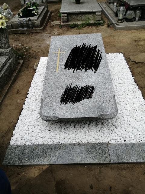 Granitowy pomnik z wygrawerowanym krzyżem, otoczony białymi kamykami, na cmentarzu.