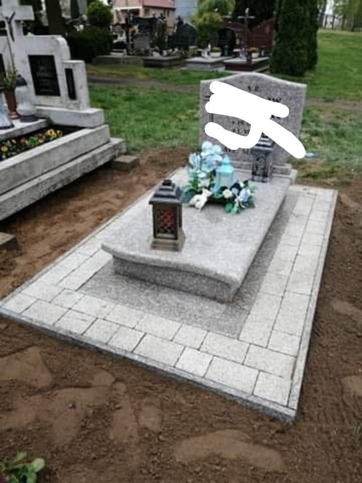 Granitowy nagrobek z lampionami i kwiatami, otoczony jasną kostką brukową na cmentarzu, ziemia wokół.