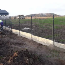 Dwóch mężczyzn pracuje przy montażu ogrodzenia z siatki na betonowej podmurówce. Widoczny fragment ukończonego ogrodzenia, teren budowy, budynek gospodarczy w tle.