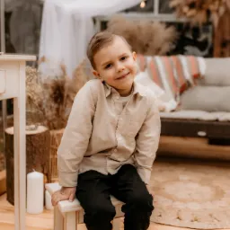 Uśmiechnięty chłopiec w beżowej koszuli siedzi na małym białym krzesełku w jasnym wnętrzu z dekoracjami z suszonych traw.