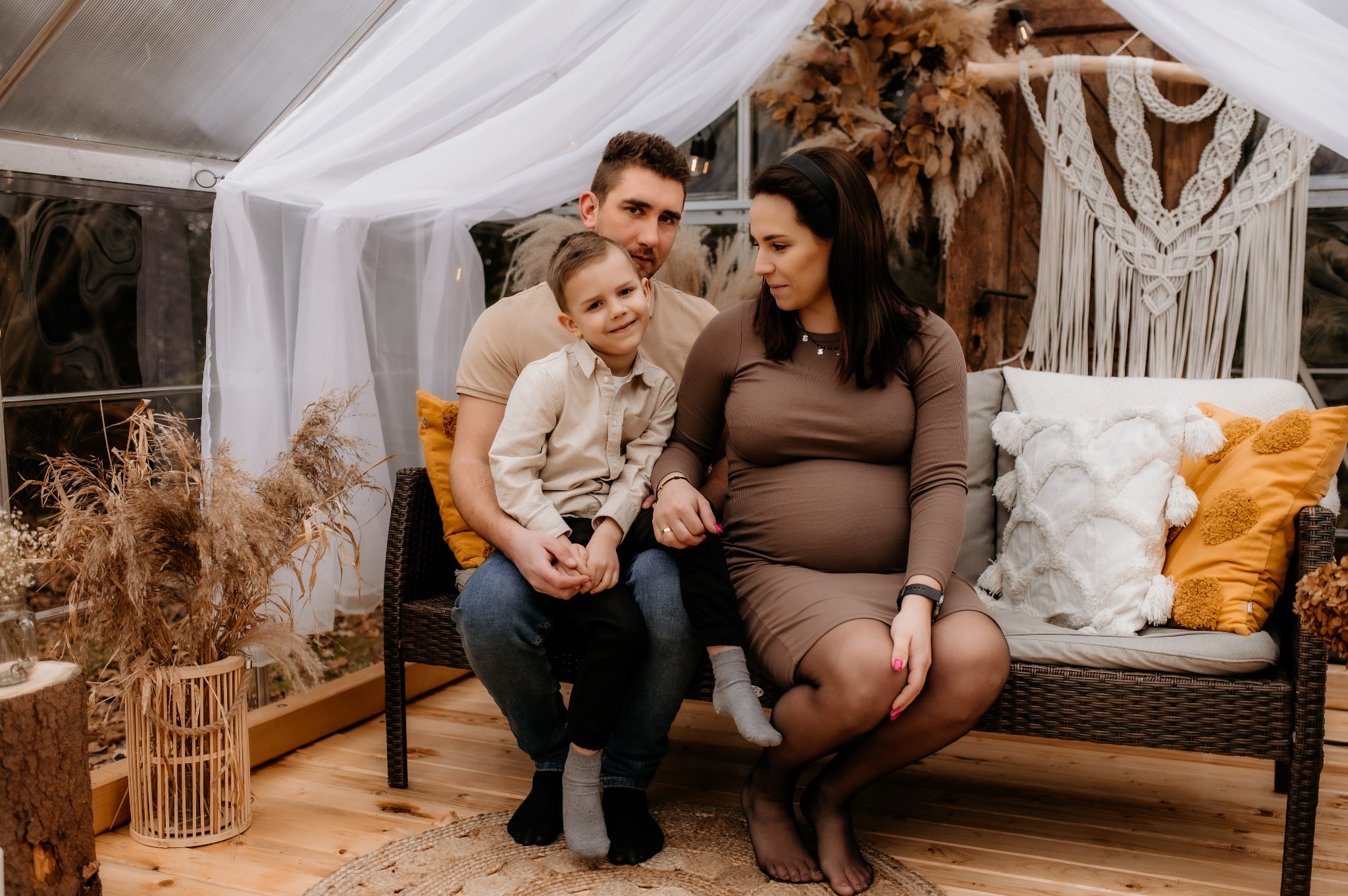 Szczęśliwa rodzina: tata trzyma synka na kolanach, obok siedzi uśmiechnięta mama w ciąży, w tle dekoracje w stylu boho i naturalne światło.