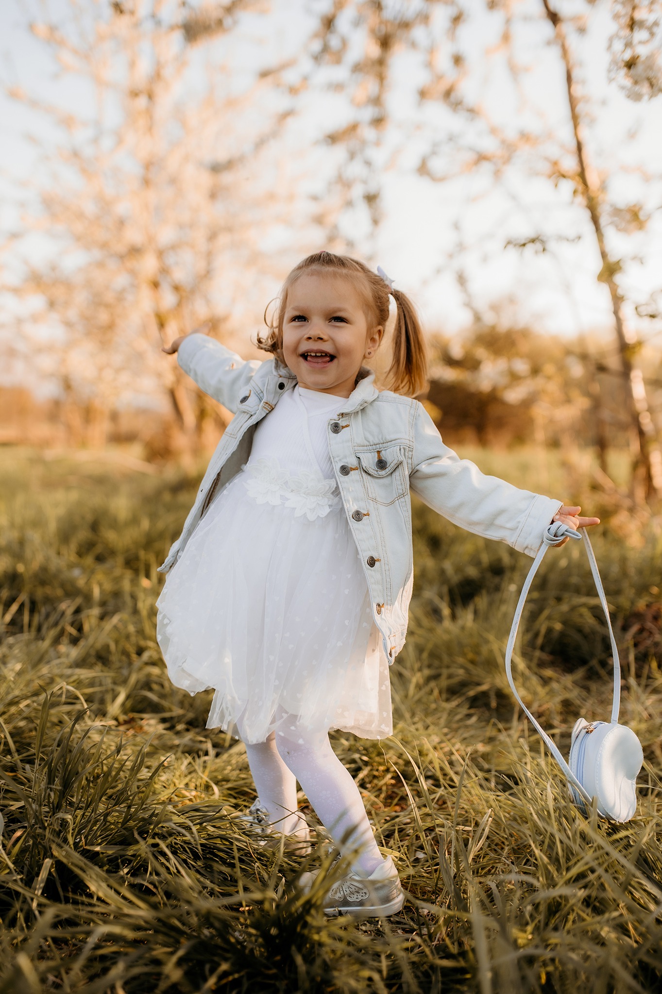 Radosna dziewczynka w białej sukience i dżinsowej kurtce, z małą torebką w kształcie serca, bawi się na łące w Mizerowie, w tle kwitnące drzewa.