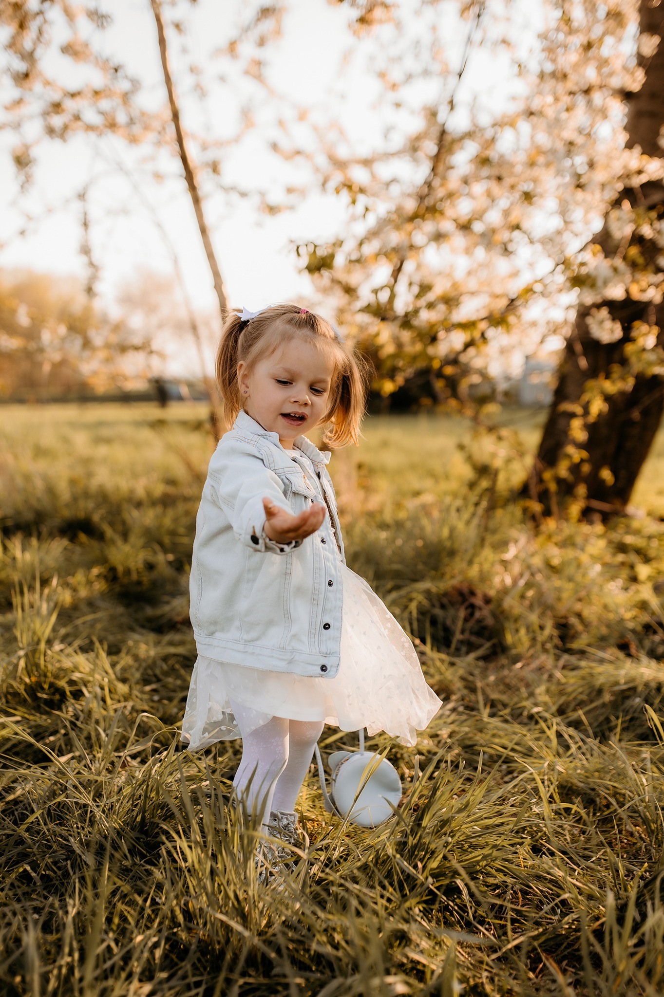 Mała dziewczynka w białej sukience i jeansowej kurtce stoi na łące, wyciągając dłoń, z drzewem w tle.
