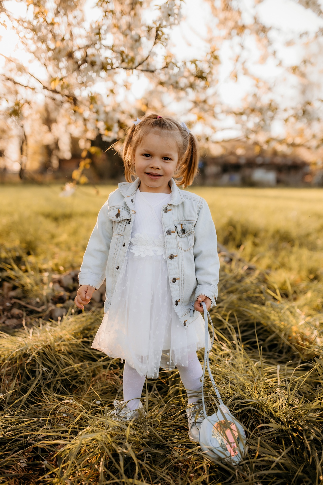 Uśmiechnięta dziewczynka w białej sukience i dżinsowej kurtce, stojąca na trawie przed kwitnącym drzewem, trzymając małą torebkę.
