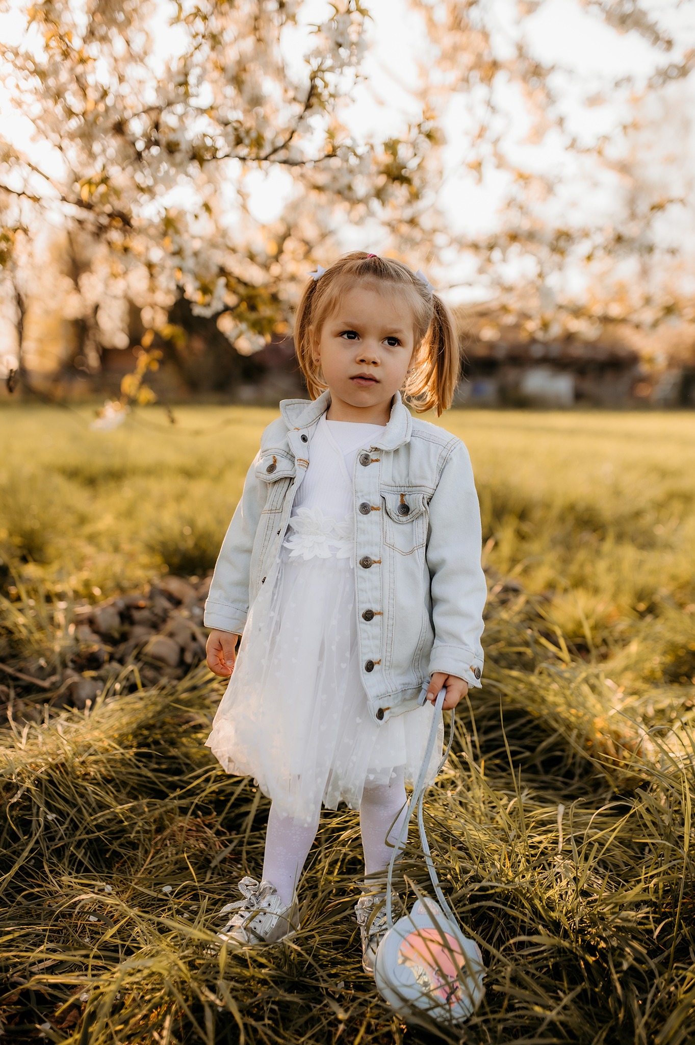 Dziewczynka w białej sukience i jeansowej kurtce stoi na łące, trzymając torebkę, z kwitnącym drzewem w tle.