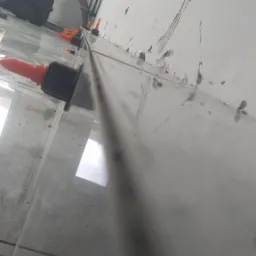 Pracownik na kolanach, poziomuje szare płytki podłogowe z użyciem pomarańczowych klinów i czarnych dystansów, widoczne zabrudzenia na ścianie.