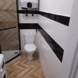 Wnętrze łazienki z toaletą, bojlerem i kabiną prysznicową, wykończone płytkami w biało-czarne pasy oraz drewnopodobnymi płytkami ułożonymi w jodełkę na podłodze.