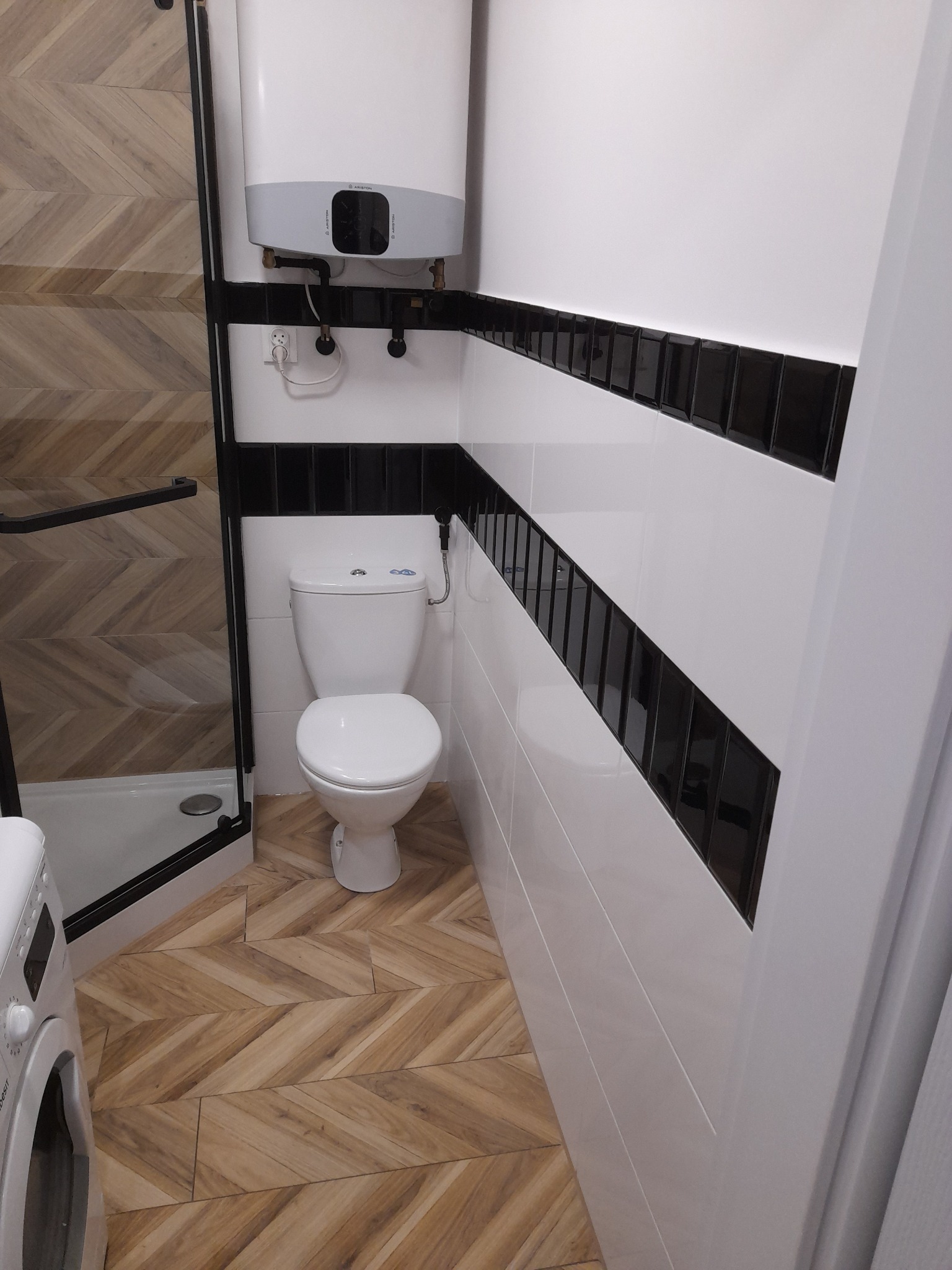 Wnętrze łazienki z toaletą, bojlerem i kabiną prysznicową, wykończone płytkami w biało-czarne pasy oraz drewnopodobnymi płytkami ułożonymi w jodełkę na podłodze.