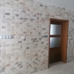 Ściana wyłożona dekoracyjną cegłą w odcieniach bieli, szarości i beżu, z widocznymi włącznikami światła i drewnianymi drzwiami z szybą w tle.