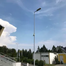 Wysoki słup oświetleniowy LED na tle błękitnego nieba z chmurami, widoczny obok budynku przemysłowego z żółtym kontenerem i schodami.