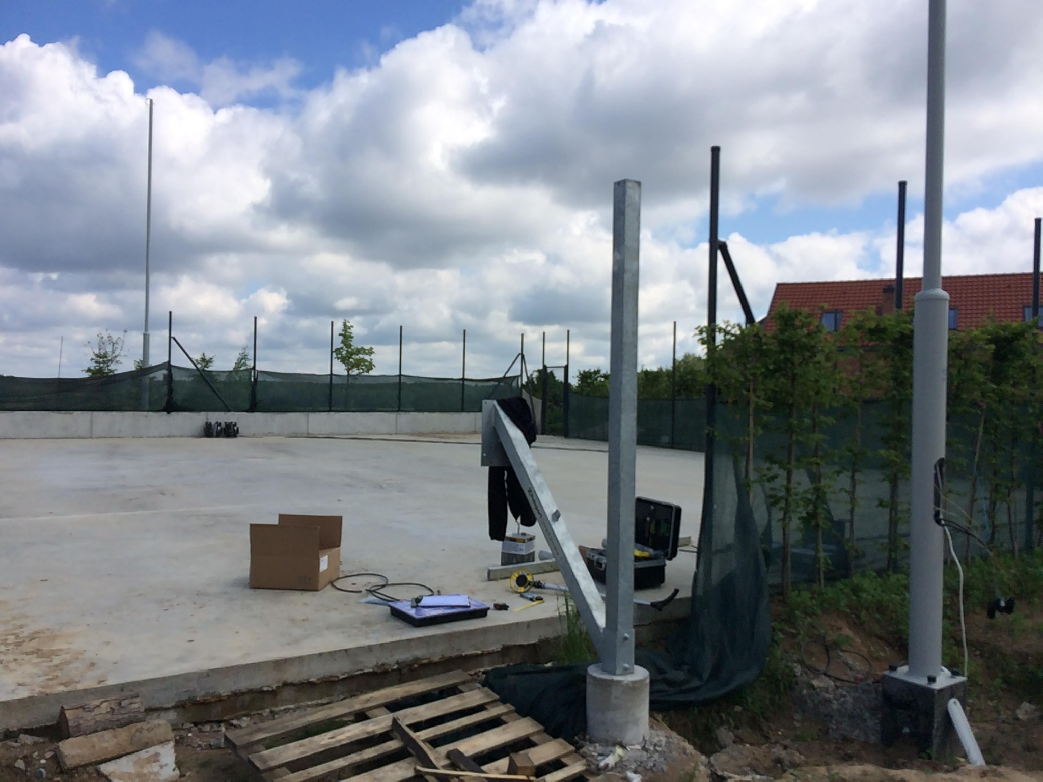 Budowa oświetlenia kortu tenisowego: widoczne słupy oświetleniowe w trakcie montażu, skrzynka narzędziowa, paleta i karton na surowym betonowym podłożu, siatka kortu w tle.
