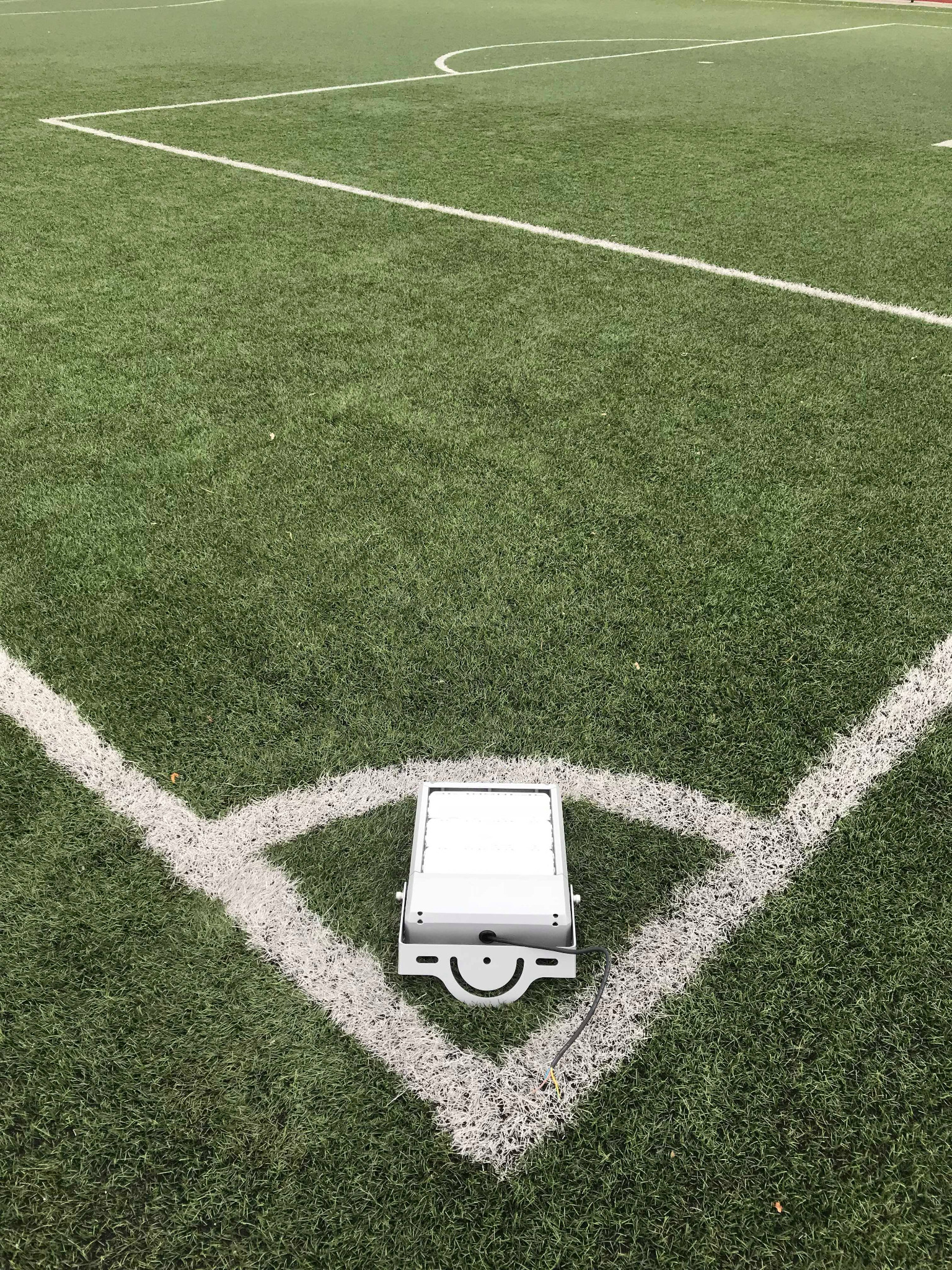 Nowa, biała lampa LED zamontowana na rogu boiska do piłki nożnej z zieloną sztuczną trawą i białymi liniami wyznaczającymi pole gry.