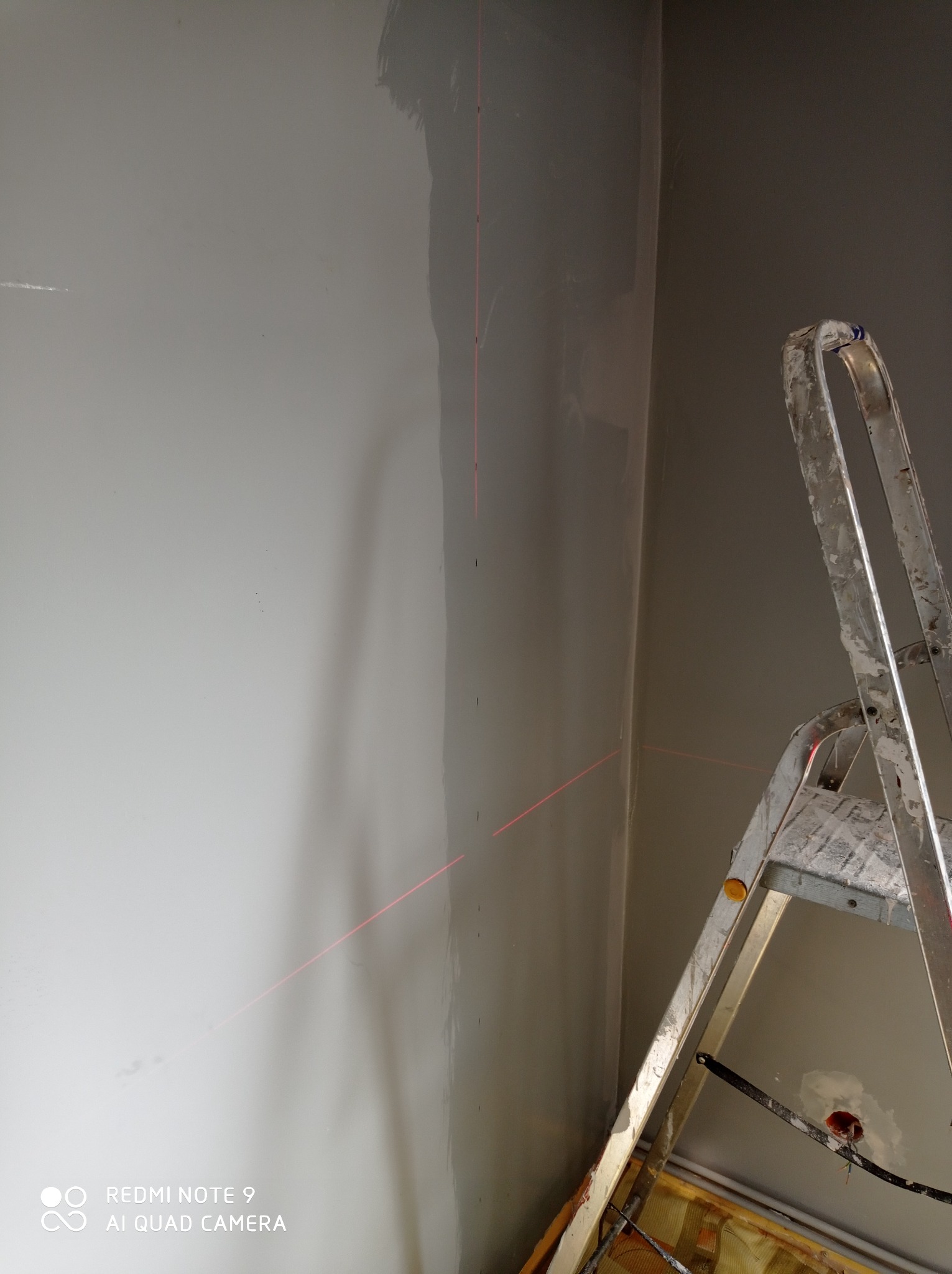 Świeżo pomalowana ściana w szarym odcieniu, widoczny laser krzyżowy wyznaczający pion i poziom, drabina malarska z resztkami farby stoi w rogu pomieszczenia.