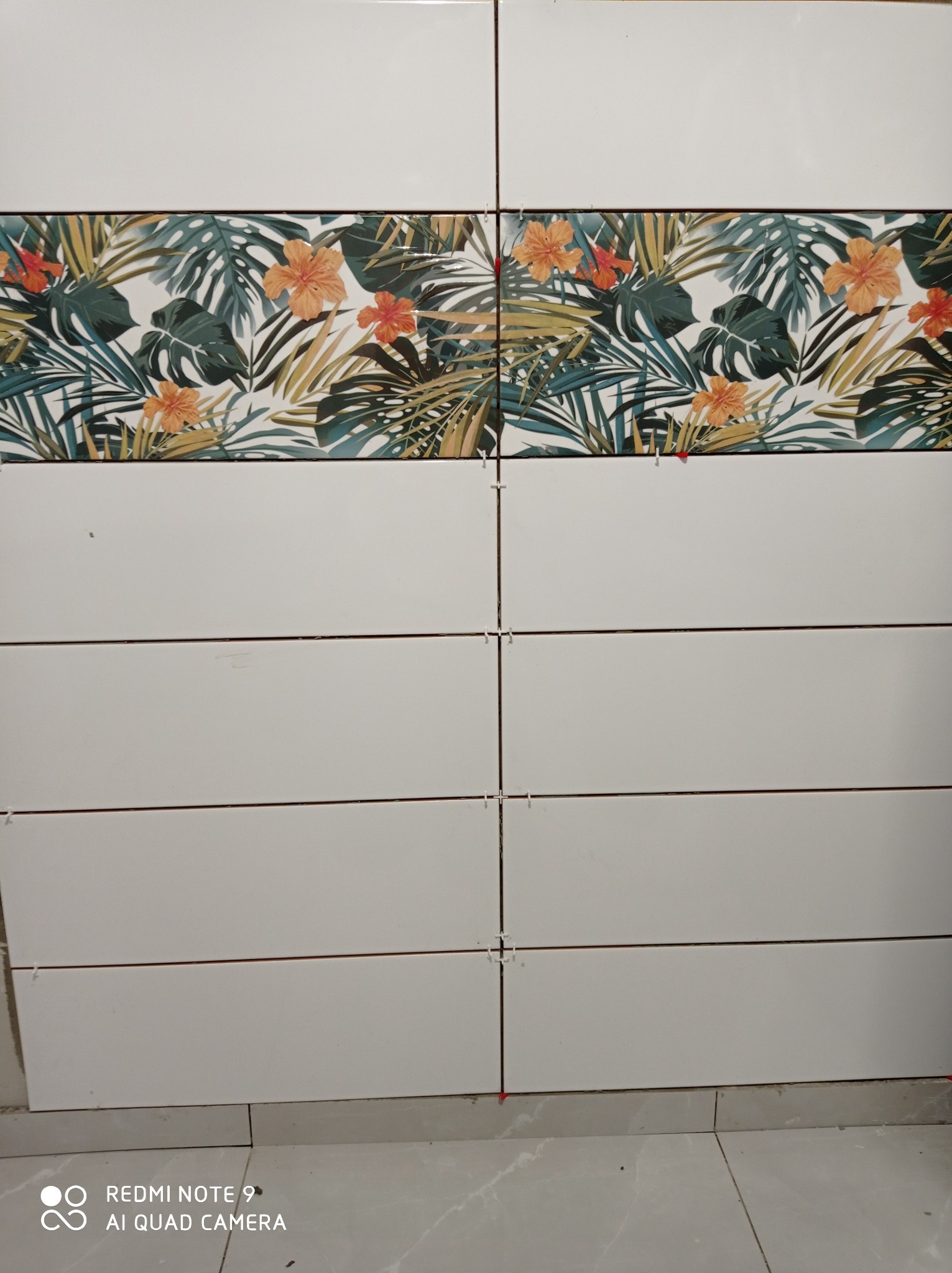 Ściana wyłożona białymi, prostokątnymi płytkami z poziomym pasem płytek dekoracyjnych z motywem tropikalnych liści i pomarańczowych kwiatów; widoczne krzyżyki dystansowe.
