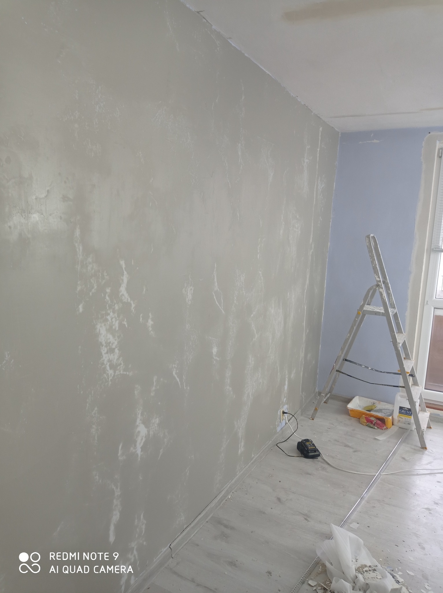 Ściana w pokoju po nałożeniu gładzi z widoczną fakturą, drabina aluminiowa, wiadro z gruntem, listwa przypodłogowa i narzędzia na podłodze.