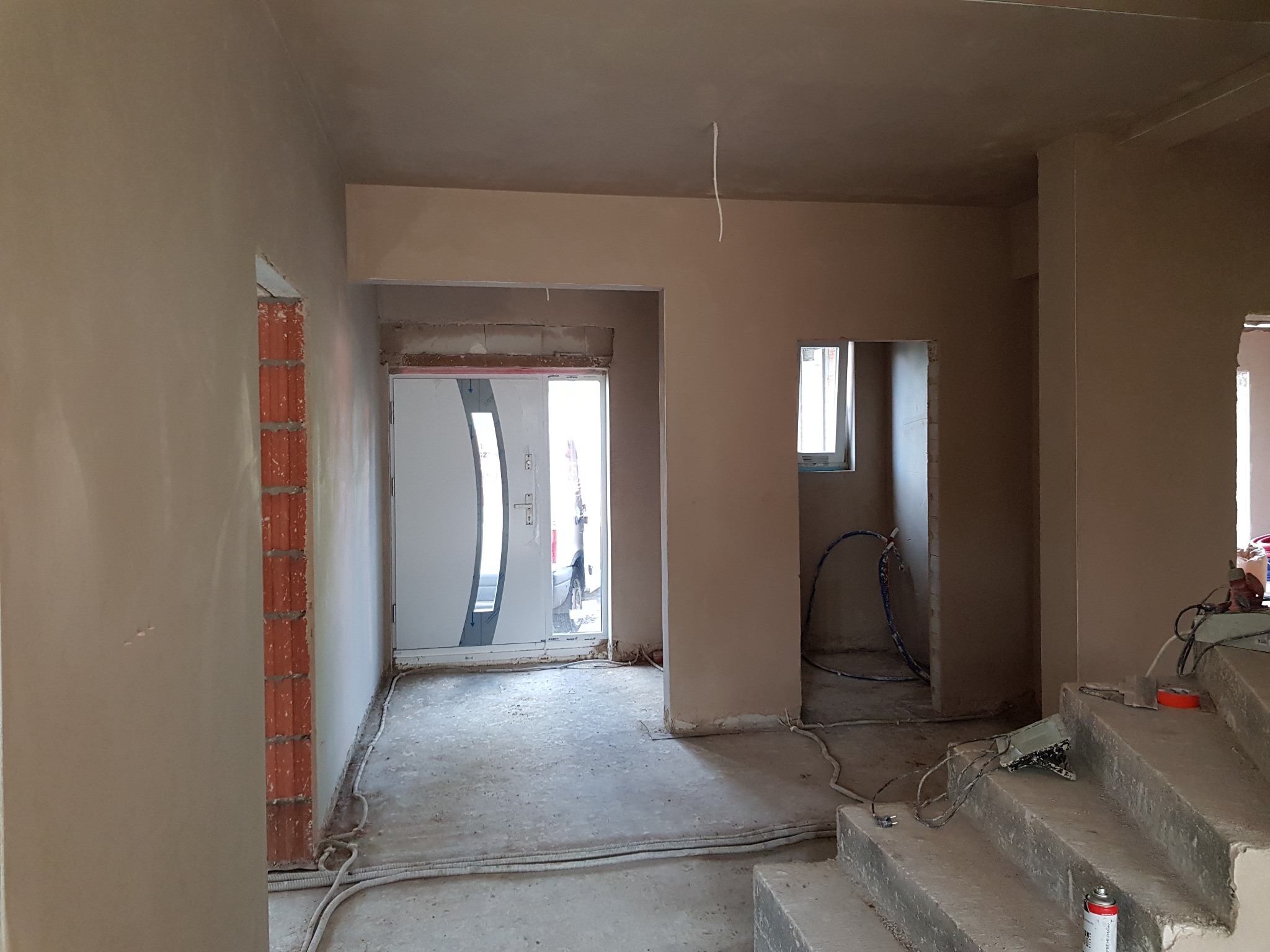 Wnętrze domu po nałożeniu gładzi gipsowej, widoczne ściany i sufit z świeżo nałożoną warstwą, niezabezpieczone kable elektryczne zwisają z sufitu, drzwi wejściowe w tle.