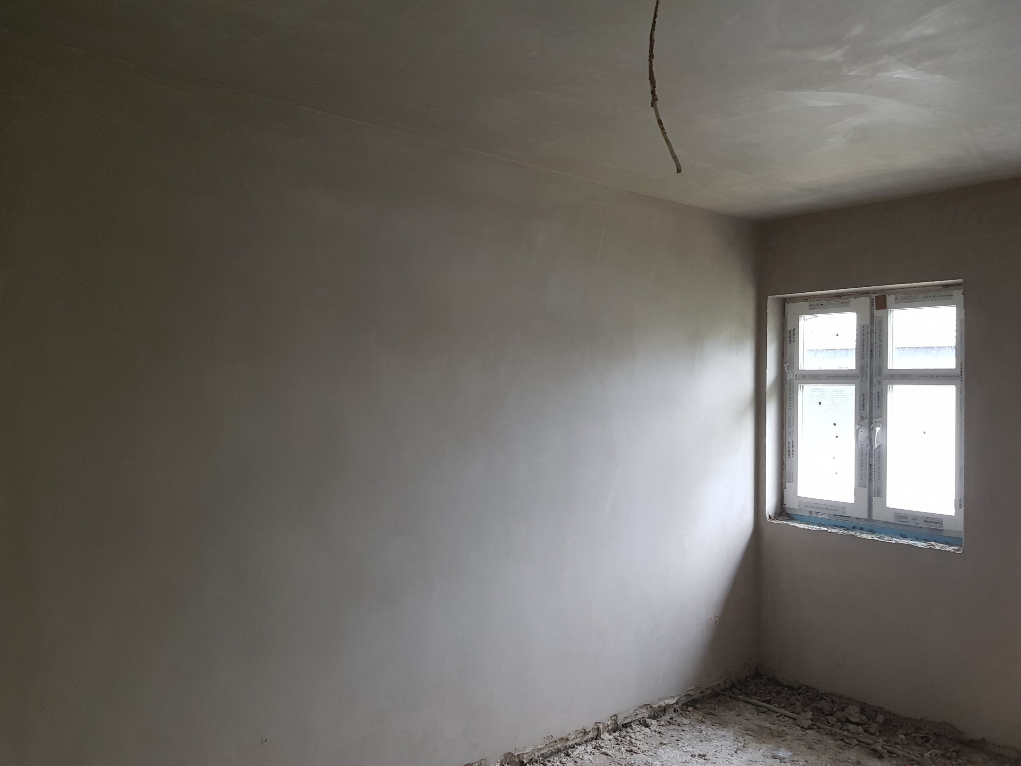 Pokój po nałożeniu gładzi gipsowej, widoczne białe okno, surowa podłoga i wystający kabel ze ściany.