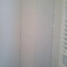 Ściana wyłożona białymi, kwadratowymi płytkami ceramicznymi w rogu łazienki, widoczny fragment białego grzejnika drabinkowego.