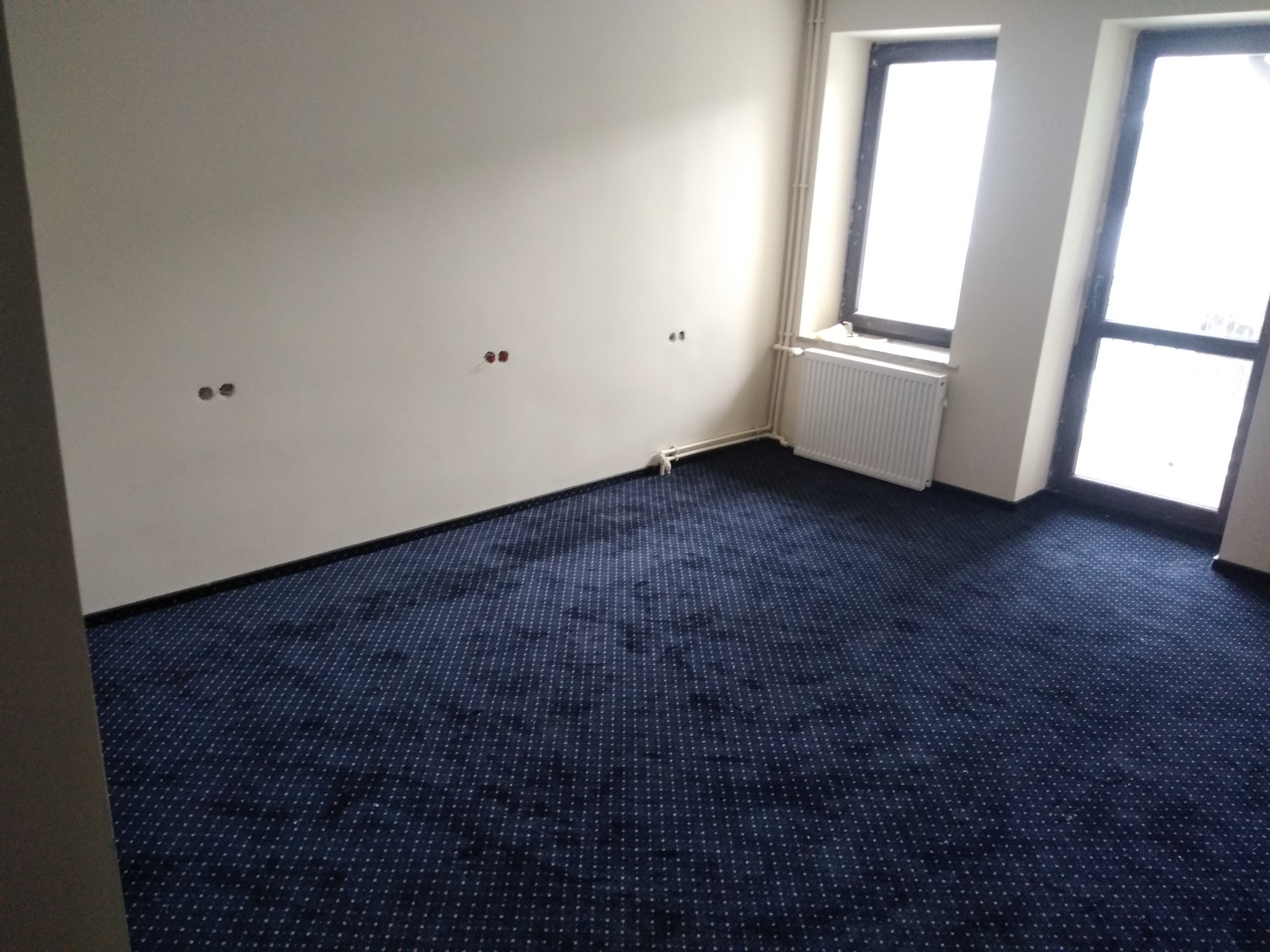 Pokój z nową, ciemnoniebieską wykładziną dywanową w drobny wzór, jasne ściany z widocznymi gniazdkami elektrycznymi, grzejnik pod oknem.