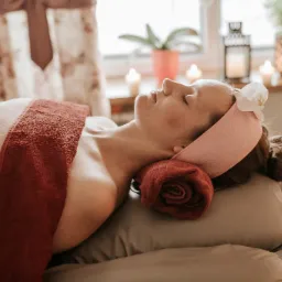 Kobieta relaksuje się podczas sesji z podgrzewanym ręcznikiem, w tle świece i rośliny doniczkowe.