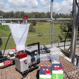 CONCRETE - Sprzęt do aplikacji preparatów, w tym maszyna z lejkiem, pistolet natryskowy i dwa pojemniki Koster, ustawione na metalowej kratownicy na tle zielonego krajobrazu.