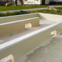 CONCRETE - Betonowa konstrukcja z niskimi ściankami działowymi, świeżo pomalowana na beżowo z białymi krawędziami, widoczna na tle budynku i zieleni.