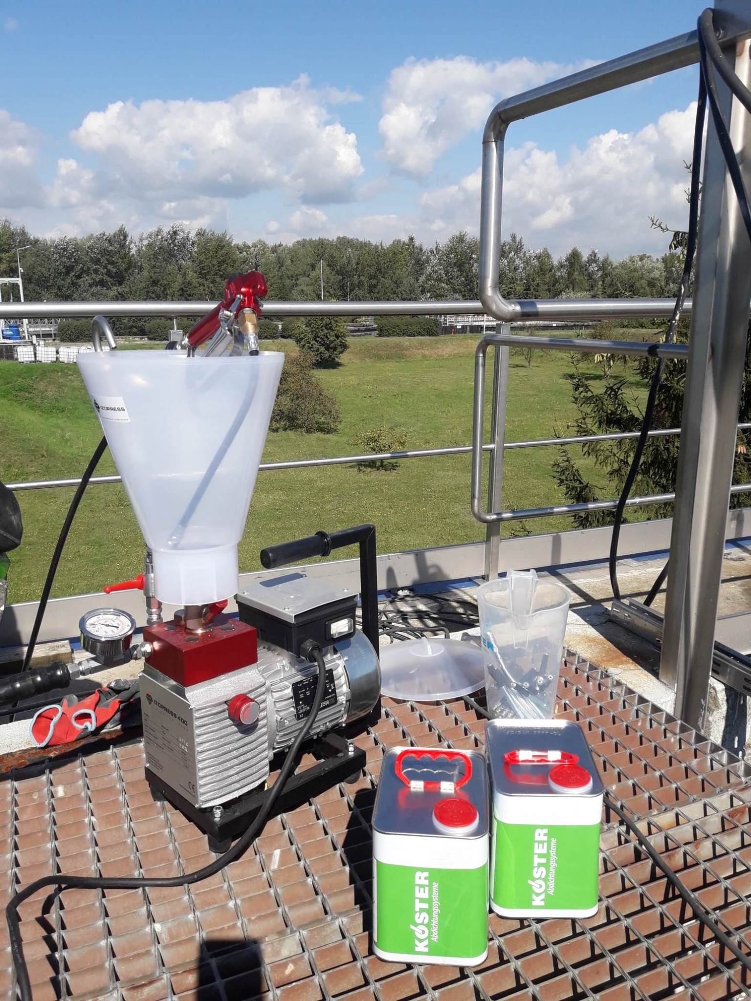 Sprzęt do aplikacji preparatów, w tym maszyna z lejkiem, pistolet natryskowy i dwa pojemniki Koster, ustawione na metalowej kratownicy na tle zielonego krajobrazu.